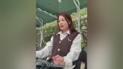 Media: Video of an Asian woman with long, straight, dark hair, wearing a white blouse and brown vest, seated outdoors under a green canopy, holding a smartphone, surrounded by greenery.