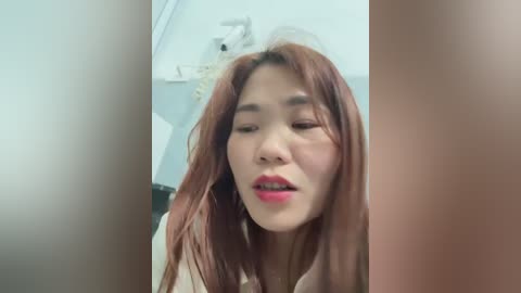 Media: Video of an Asian woman with long, straight auburn hair, wearing red lipstick, looking upward with a neutral expression. Background features a white wall with a white door partially visible.