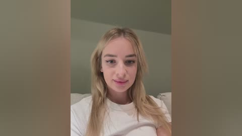 Media: Video of a young Caucasian woman with long blonde hair, fair skin, and light makeup. She wears a white t-shirt, sitting in a dimly lit room with neutral-colored walls.