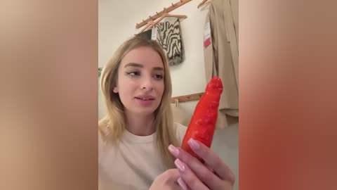 Media: Video of a young blonde woman with fair skin, holding a large, bright red dildo, in a bathroom with beige walls, zebra-patterned towel, and beige robe hanging on a hook.