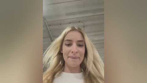 Media: Video of a young blonde woman with long hair, wearing a white shirt, taken from below, showing her smiling and looking down. Background features a metallic ceiling with visible beams and pipes.