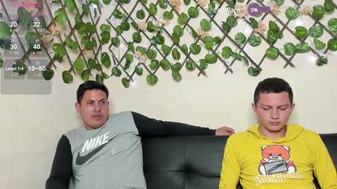 Media: Video of two men, one in a grey Nike shirt, the other in a yellow shirt with a cartoon fox, sitting on a black couch against a wall adorned with fake vines and a height chart.