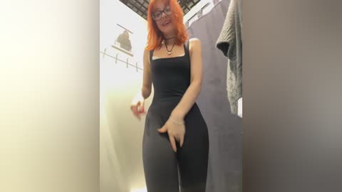 Media: A video of a slim, fair-skinned woman with red hair and glasses, wearing a tight black bodysuit, standing in a dimly lit changing room.