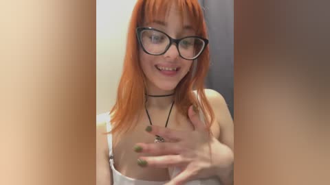 Media: Video of a young woman with straight, orange hair, wearing glasses, a black choker, and a white tank top, smiling, with blurred figures in the background.