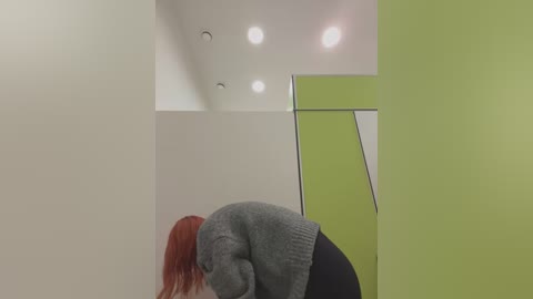 Media: Video of a woman with long red hair, wearing a grey sweater and black pants, bending over in a minimalist room with lime green and white walls, three recessed ceiling lights, and a reflective mirror.