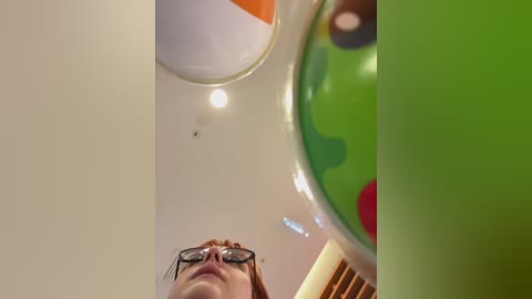 Media: A video shows a person with short, red hair and glasses looking up at a colorful, round ball with green, red, and black spots, set against a white ceiling.