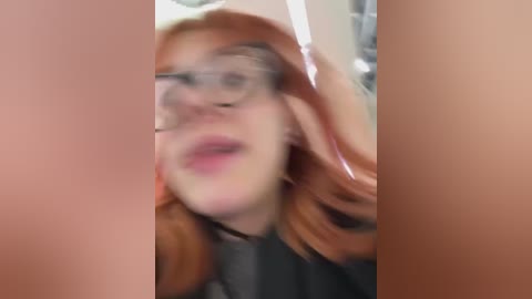 Media: Video of a young woman with pale skin and long, wavy red hair, wearing glasses, and a black top. She is looking directly at the camera with an expression of surprise or shock.