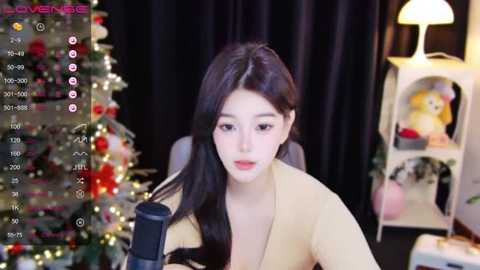 Media: Video of an East Asian woman with long black hair, fair skin, wearing a beige top, sitting in a dimly-lit room with a Christmas tree and a shelf with decorations. She's holding a microphone.