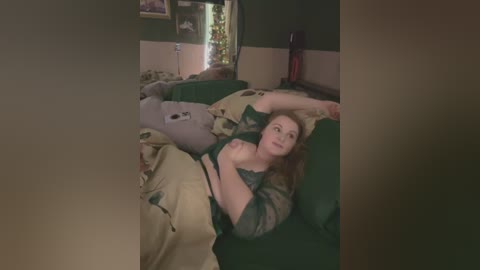 Media: Video of a young woman with long hair, lying on a green couch in a dimly lit room with green and beige walls, wearing a green and black patterned top, partially covered by a blanket.