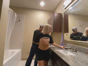 Media: Video of a blonde woman in a black crop top and shorts, washing her hands in a beige-tiled bathroom. A man in a black cap and dark clothes is behind her, observing.