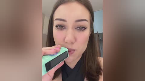 Media: Video of a young woman with fair skin, brown hair, and sharp makeup, including winged eyeliner, using a green phone to take a selfie in a modern bathroom with a white bathtub and towel rack.