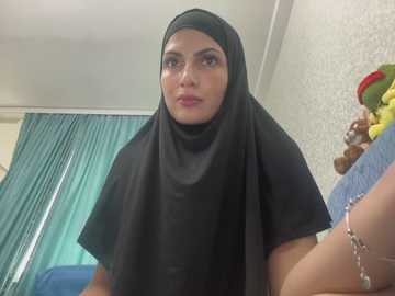 Media: Video of a middle-aged woman with a fair complexion, wearing a black hijab and short-sleeved shirt, standing in a room with teal curtains and floral wallpaper.