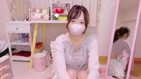 Media: Video of an Asian woman in a pink face mask, light gray hoodie, and white pants, sitting on a pink rug in a cluttered, pastel-themed room with a white shelf, mirror, and pink chair.