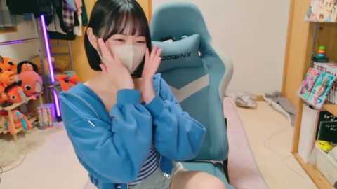 Media: Video of an Asian woman with short black hair in a blue hoodie, wearing a face mask, sitting in a blue gaming chair in a cluttered room with plush toys and shelves.