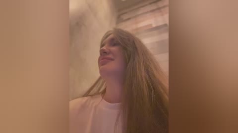 Media: Video of a young woman with long brown hair, wearing a white top, smiling gently, standing in a dimly lit, concrete hallway with a window.