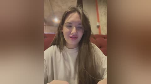 Media: Video of a young woman with long brown hair, wearing a beige top, sitting in a red cushioned booth with a blurred background.