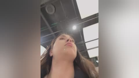 Video of a young woman with fair skin and long brown hair, looking upward with a serene expression. The background features a modern, industrial-style ceiling with exposed beams and lights.