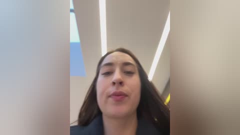 Media: Video of a young woman with fair skin and long dark hair, making a duck face, standing in a modern, brightly lit interior with angular ceiling panels and recessed lights.