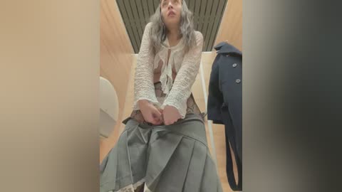 Media: A video of a woman with shoulder-length grey hair, wearing a lace top and grey skirt, seated on a toilet stall bench, with a navy coat hanging nearby.