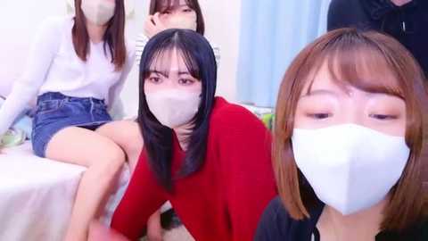 Media: Video of four Asian women in casual clothing, including a red shirt, sitting closely, wearing face masks, with one smiling, in a brightly lit room.