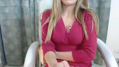 Media: Video of a fair-skinned woman with long blonde hair, wearing a red V-neck sweater, sitting on a white chair with arms crossed. Background features gray and white patterned curtains.