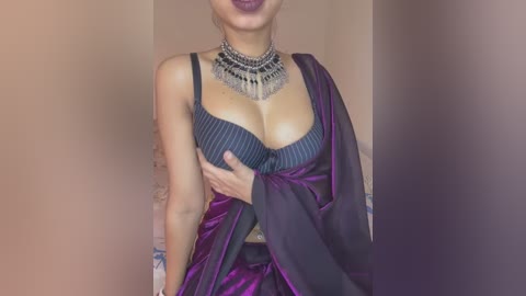 Media: Video of a South Asian woman in a dark purple sari, revealing her cleavage in a black and white striped bra. She wears a silver choker necklace. Background is a beige wall.