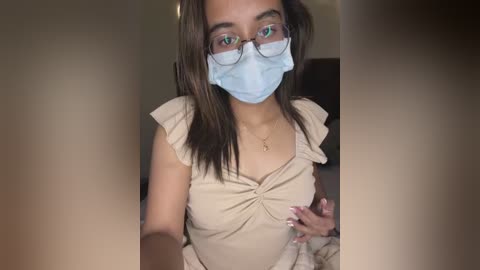 Media: A video of a young woman with medium-brown skin, wearing a light beige top, blue face mask, and glasses, holding a phone.