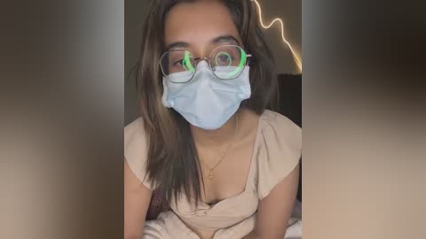 Media: Video of a young woman with medium brown skin, wearing a light blue surgical mask, round glasses, and a beige blouse, sitting indoors with blurred background.