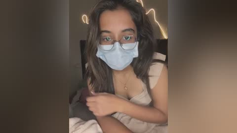 Media: A video of a young woman with medium-brown skin, long dark hair, and glasses, wearing a blue mask and a light-colored top. She is indoors, sitting on a bed with a white pillow, with warm, soft lighting.