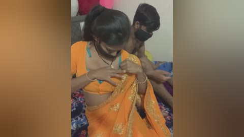 Media: Video of a young Indian woman in an orange saree with gold embroidery, adjusting her top while a man with a black mask watches, in a dimly lit room.