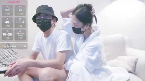 Media: A video of a young Asian couple in casual white attire, seated on a bed with a digital thermometer showing 10.5\u00b0C, wearing face masks in a minimalist bedroom.