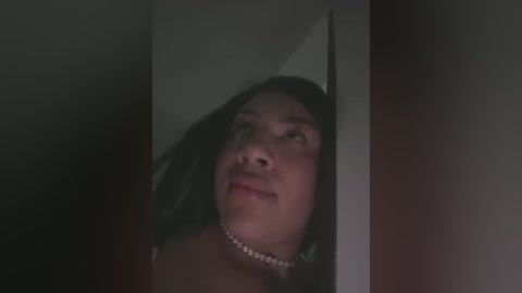 Media: Video of a young woman with medium brown skin and long, straight black hair, looking up with a contemplative expression, framed by a white door. She wears a silver choker necklace.