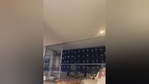 A video of a modern living room with floor-to-ceiling windows revealing a night sky dotted with twinkling lights. The room features a beige sofa and a wooden coffee table, with a cozy, minimalistic ambiance.