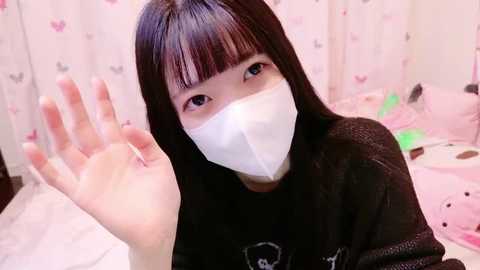 Media: A video of an East Asian woman with straight black hair and a white surgical mask, waving, indoors with a pink and white themed room in the background.