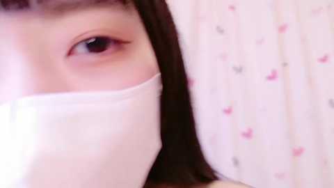 Media: Video of a young Asian woman with light skin, dark hair, and hazel eyes, wearing a white surgical mask, lying on a bed with a pink floral-patterned pillow, giving a close-up view of her face.