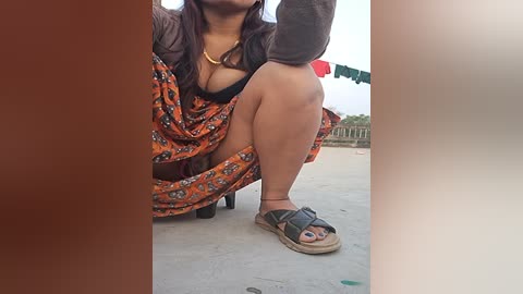 Media: Video of a woman with medium skin tone squatting on a balcony. She wears an orange floral dress, black bra, and beige sandals, with a gold necklace. Background includes red traffic lights and blurred greenery.