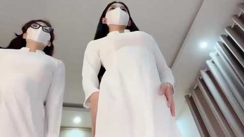 Media: Video of two women in white lab coats and face masks, standing indoors, one wearing glasses, the other with long black hair, in a modern, well-lit room with grey curtains.