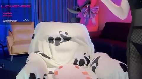 Video of a person in a white cow-print robe, standing in a modern living room with red and blue lighting, a couch, and a wooden chair.