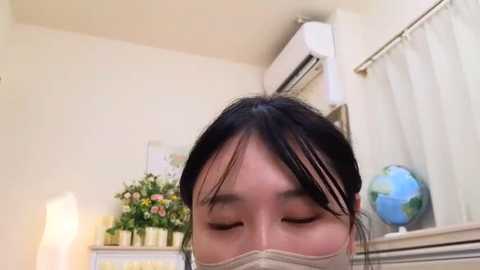 Media: Video of an Asian woman with long black hair, wearing a beige mask, closed eyes, in a minimalist room with white walls, potted plants, a globe, and an air conditioner.