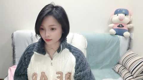 Media: Video of a young East Asian woman with short black hair, fair skin, and light makeup, wearing a gray and white fleece jacket, sitting on a couch. The background features a stuffed toy and striped pillows.