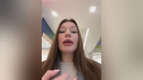 Media: A close-up photo of a young Caucasian woman with straight brown hair, fair skin, and full lips, wearing a light-colored shirt, captured from a low angle in a brightly lit room with modern decor.