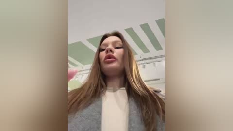 Media: Video of a fair-skinned woman with long, straight brown hair, wearing a gray cardigan over a white shirt, in a room with pastel green walls and white ceiling beams.