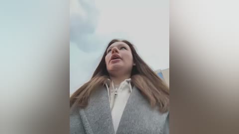 Media: A video captures a young woman with long brown hair, wearing a gray sweater and white shirt, looking upward with a slight smile, framed by soft, blurred light.