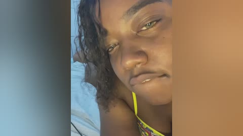 Media: Video of a young Black woman with medium brown skin, curly hair, and a piercing nose stud, lying on a bed, wearing a yellow and black patterned top.