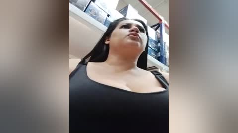 A video of a plus-sized woman with fair skin, wearing a black tank top, standing in a department store, looking upwards. The background features clothing racks and a staircase.