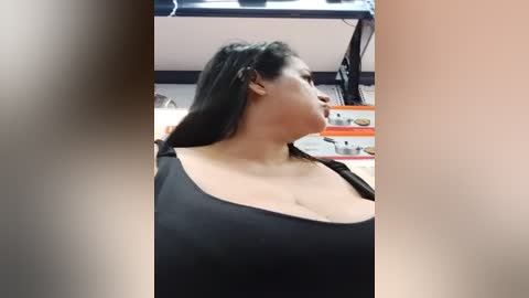 A video of a curvy, light-skinned woman with long black hair, wearing a low-cut black top, standing in a store with shelves and products in the background.