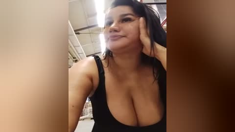 A video of a young, curvy woman with large breasts, wearing a low-cut black top, sitting in a dimly lit industrial room. She has light brown skin and long, dark hair.