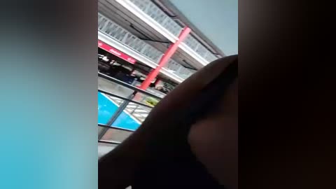 Media: Video of a blurry swimming pool scene with a red pole and a person in the foreground, partially obscured. The background features a multi-level building with white and red accents.