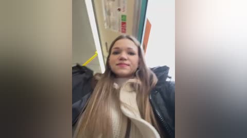 Media: Video of a young Caucasian woman with long, straight, light brown hair, wearing a beige scarf and black jacket, sitting in a subway car. Background shows other passengers and a yellow safety sign.