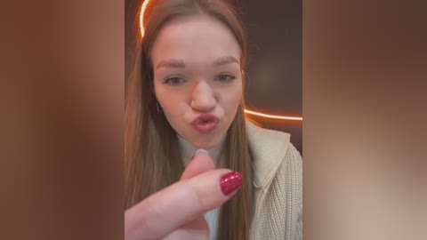 Media: A video of a young woman with fair skin, long brown hair, and a pouty expression, blowing a kiss. She wears a beige sweater and has red nail polish. The background features a blurred, warm-toned indoor setting.